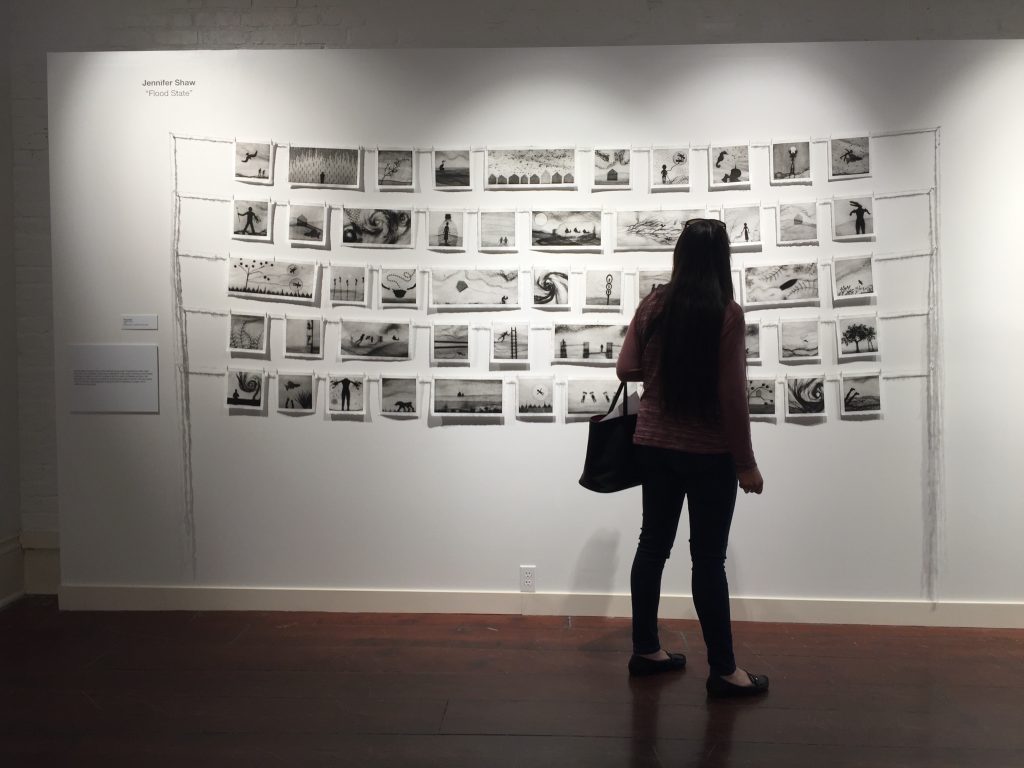 Silhouette of woman viewing Flood State installation at Louisiana State Museum. Black and white prints hang in a grid, pinned to delicate yarn.