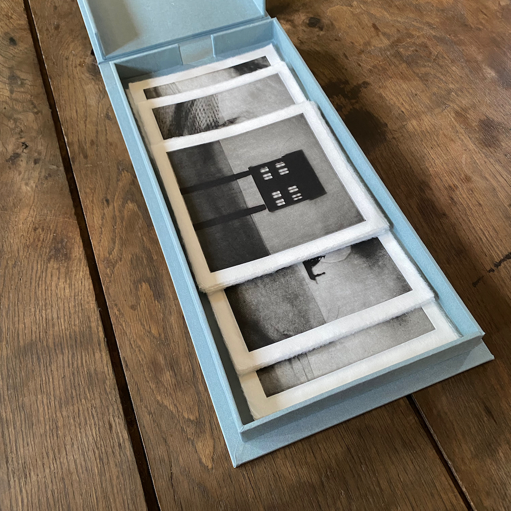 Blue clamshell portfolio, open on a wooden tabletop, with black and white photogravure prints inside.