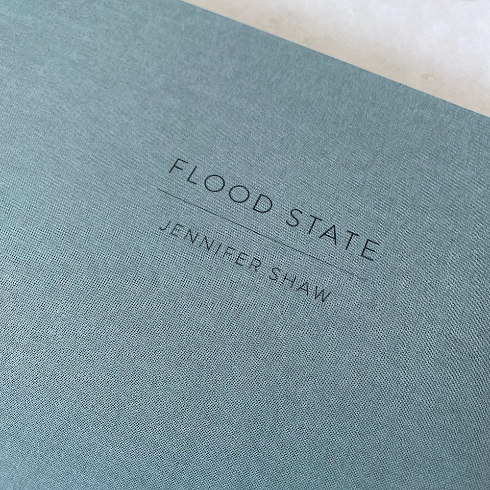 Cover detail of portfolio box, showing texture of blue cloth and title imprint which reads: FLOOD STATE - Jennifer Shaw, printed in black.